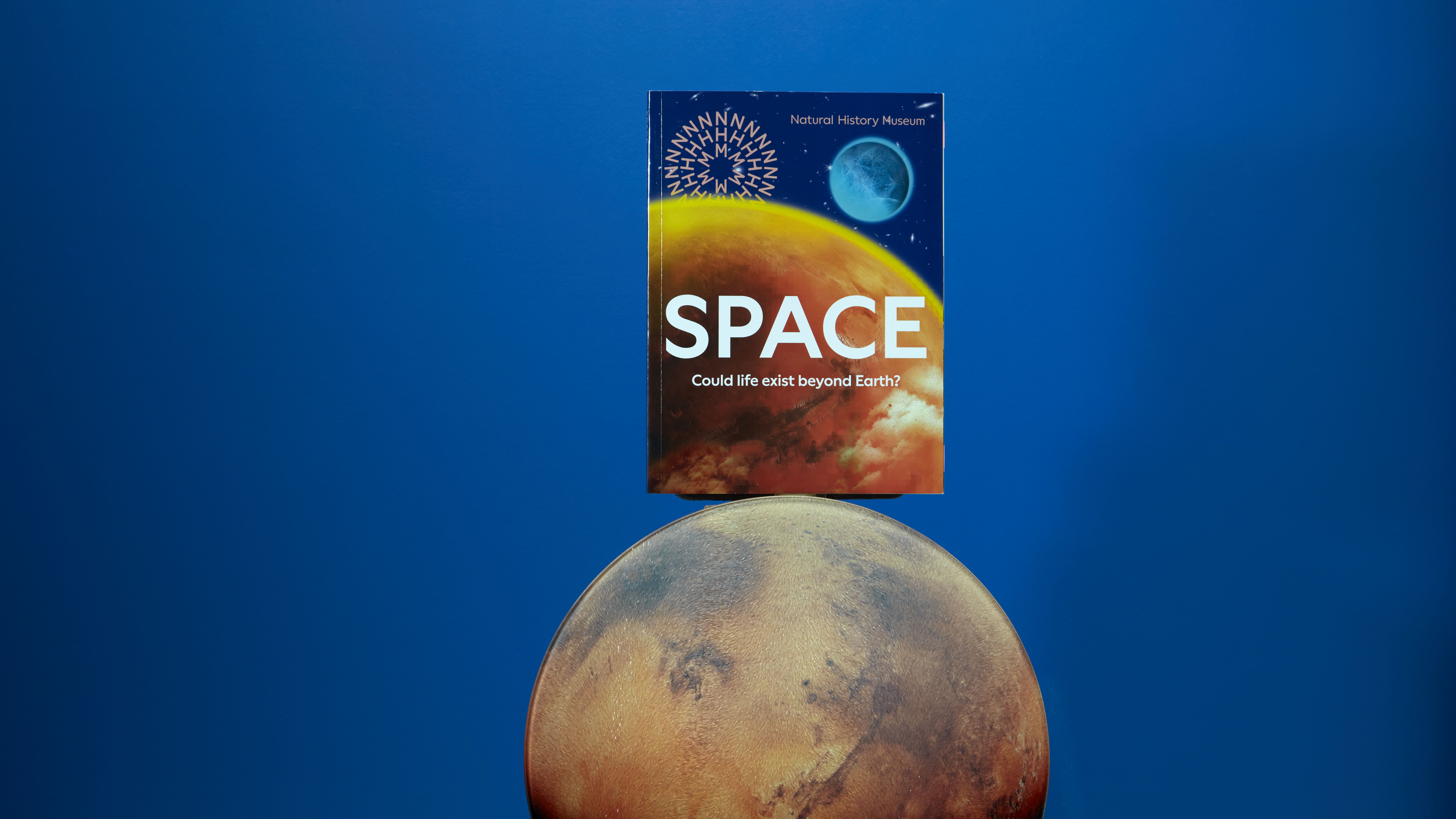 The Space exhibition book balances on top of a Mars-like planet, against a dark blue background.